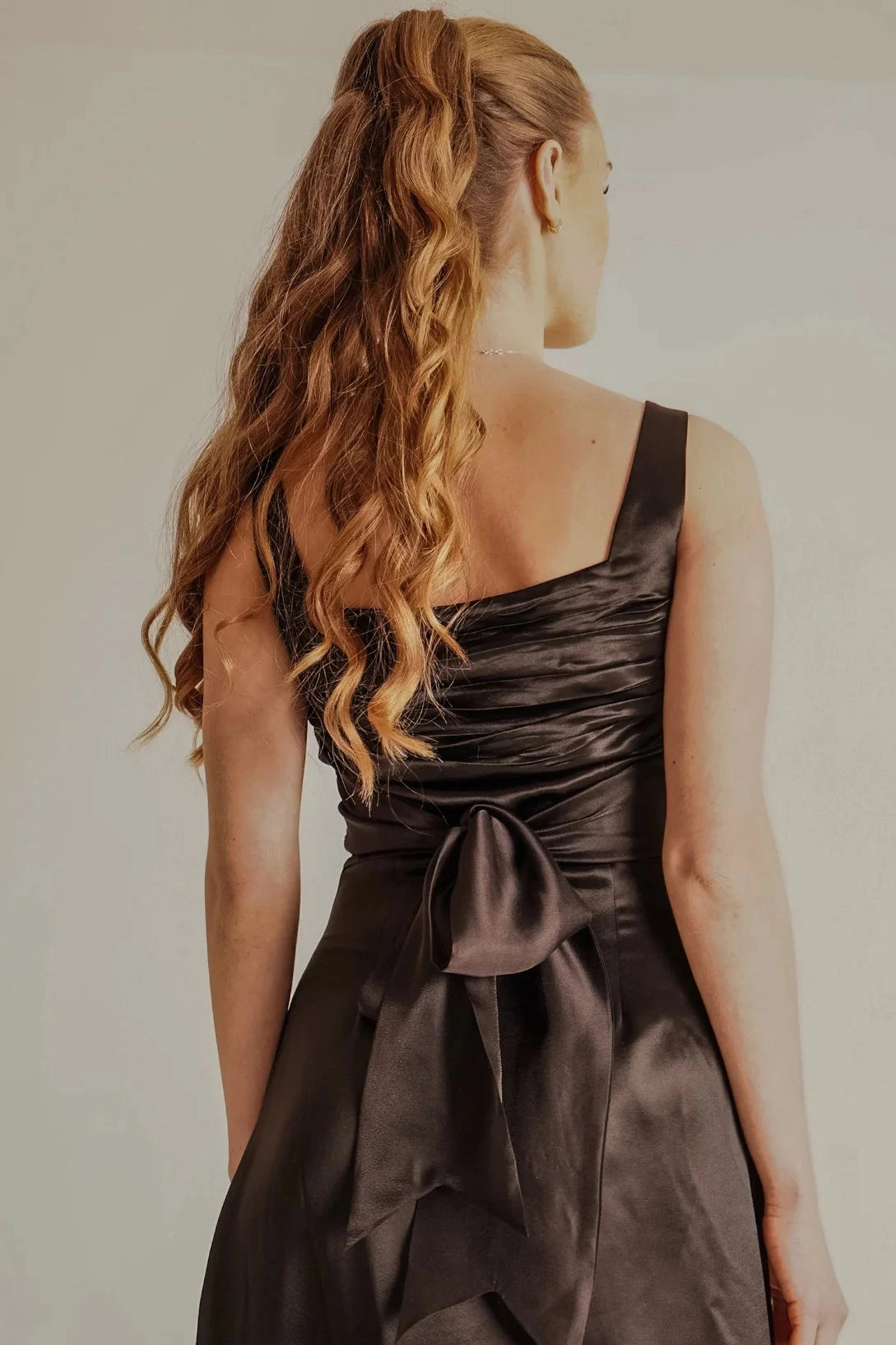 Woman wearing a dark brown satin dress with a bow detail on a plain background